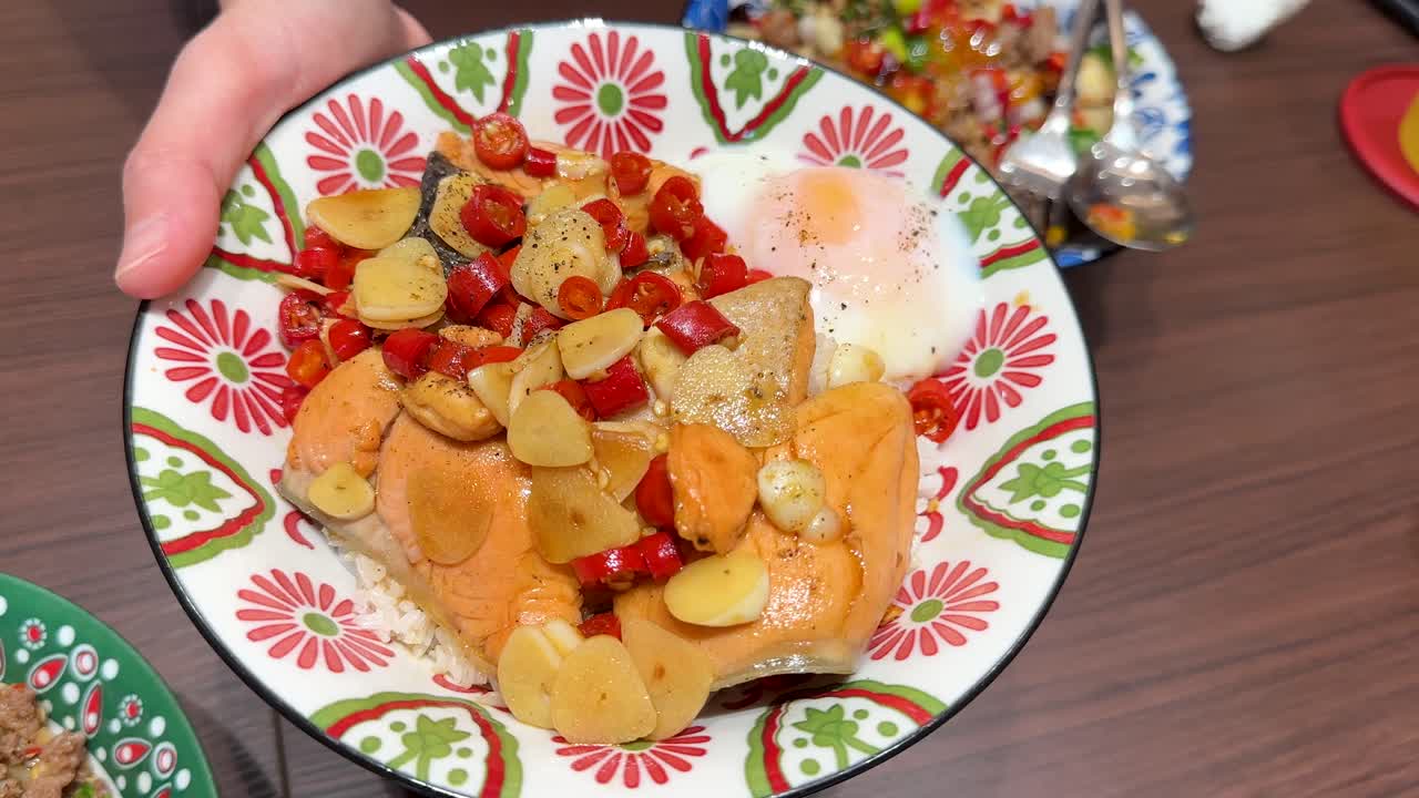 A hand presents a vibrant salmon rice bowl topped with vegetables, sauce, and a soft-cooked egg in a well-lit indoor dining setting with overhead camera movement