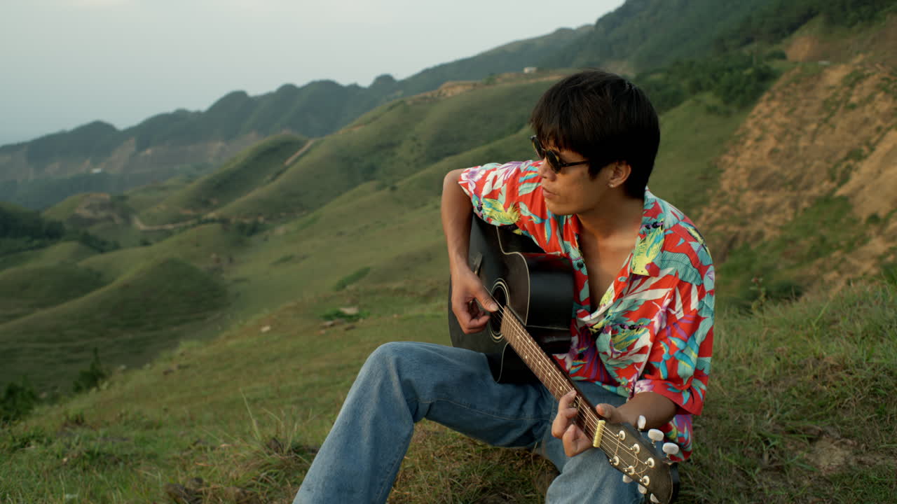 Asian Male Young Musician Plays Guitar at Green Hills Landscape, Colorful Shirt and Sunglasses