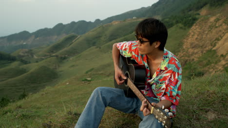 asian male young musician plays guitar at green hills landscape, colorful shirt and sunglasses