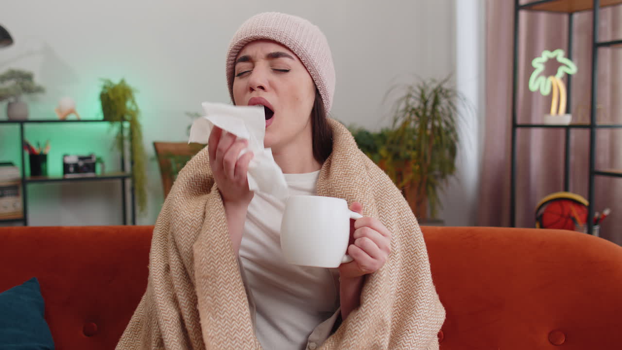 Sick ill woman suffering from cold or allergy sitting on home sofa sneezes wipes snot into napkin
