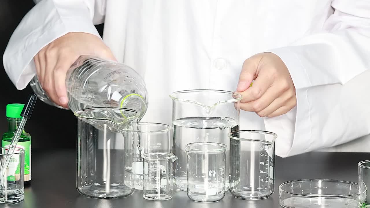 Una persona con un camice da laboratorio bianco versa dell'acqua da una bottiglia in becher di vetro