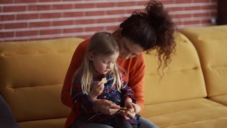 madre rizada y hija pequeña comiendo galletas en casa en el sofá