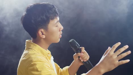 close up side view of young asian boy holding a microphone and singing on the white smoke black background