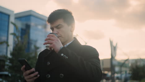 investor in black coat walking outdoors, thoughtfully browsing his phone while sipping coffee in vibrant urban setting featuring modern buildings, parked cars, greenery, and people walking