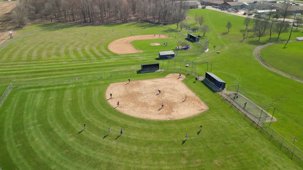 Riprese aeree con drone catturano l'azione vivace di sportivi che giocano a baseball su un vasto campo verde in Svizzera, evidenziando l'armonia tra lo sport e il paesaggio naturale.