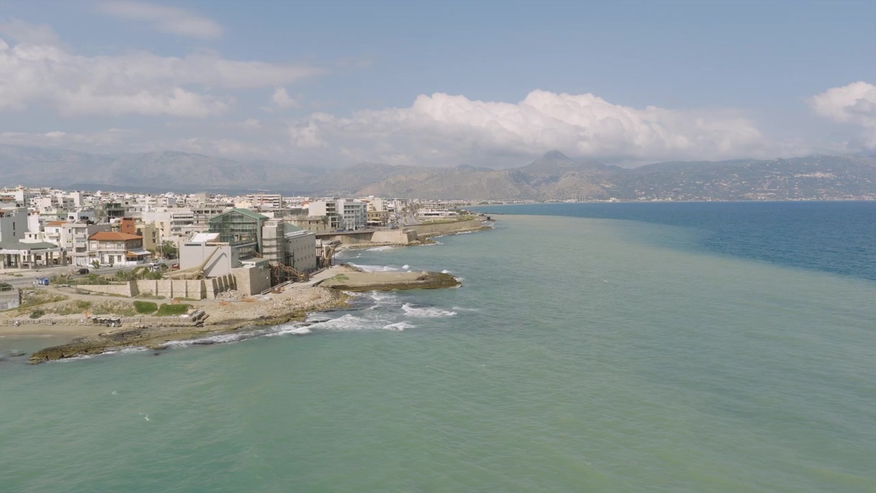 Vista aérea de la costa de Heraclión en Creta, donde las aguas poco profundas de color turquesa se encuentran con el mar azul profundo, con el borde de la ciudad y las montañas elevándose en la distancia.