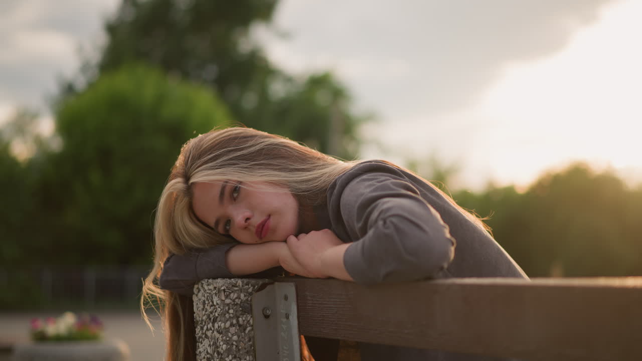 Lady seated on bench at edge, resting her head and arms with a calm expression, as sunlight creates reflective light around her, background features trees with a blurred view of people walking
