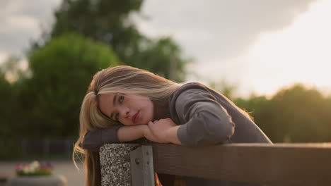 lady seated on bench at edge, resting her head and arms with a calm expression, as sunlight creates reflective light around her, background features trees with a blurred view of people walking