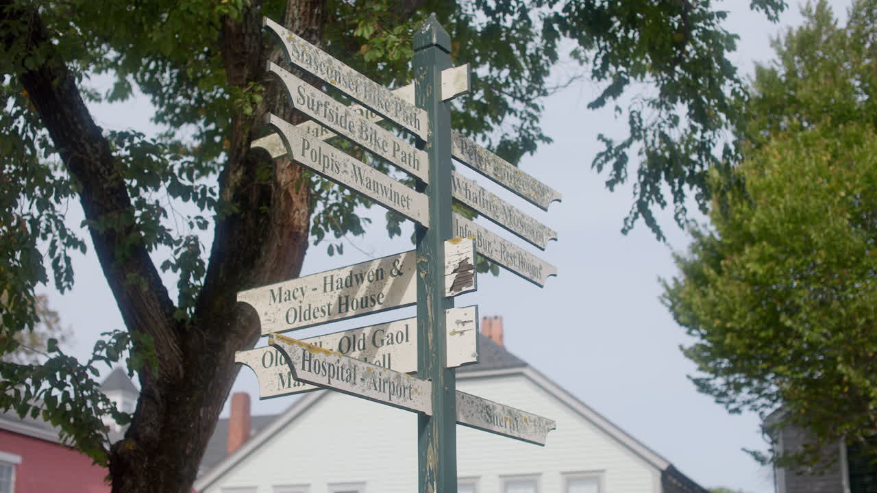Håndholdt optagelse af et vejrbidt destinationsskilt i Nantucket, Massachusetts, der peger mod lokale vartegn under løvrige træer i blødt dagslys.