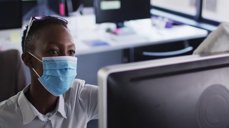 Eine afroamerikanische Frau mit Gesichtsmaske reinigt ihren Computer mit einem Tuch, während sie an ihrem Schreibtisch sitzt.
