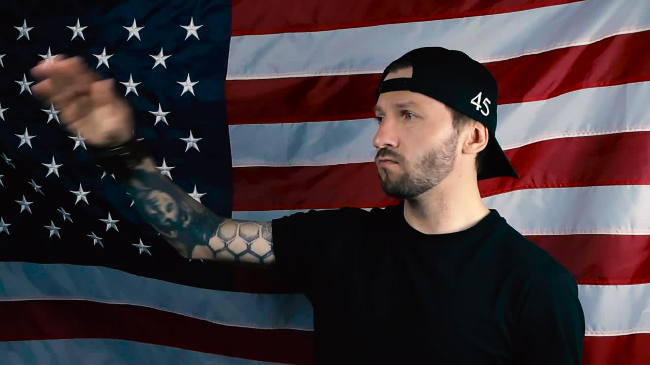 A proud patriot with tattooes and black clothing saluting in front of the Unites States of America flag in slow motion with determination and loyalty to the country, flag moving slightly in the wind.