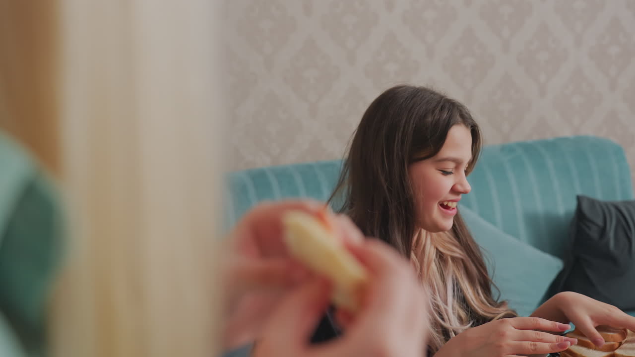 Una ragazza seduta sul divano reagisce emotivamente durante una conversazione mentre tiene del pane, il primo piano sfocato mostra una mano che tiene un sandwich, in un'ambientazione mattutina casual con interni domestici e luce naturale visibile.