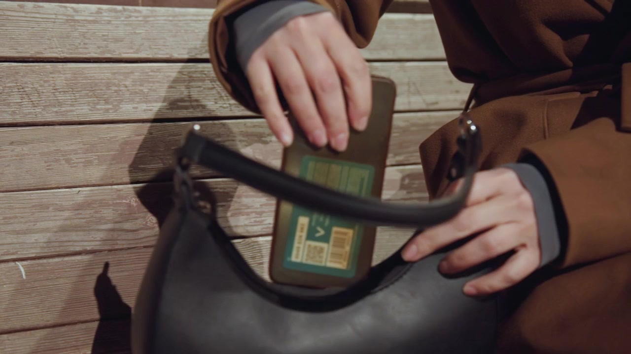 Donna su una panchina che recupera il portafoglio, scena notturna urbana ansiosa con borsa di pelle aperta, dita che cercano una carta all'interno di una tasca con cerniera, manica di cappotto marrone e mani in primo piano che rivelano tensione.