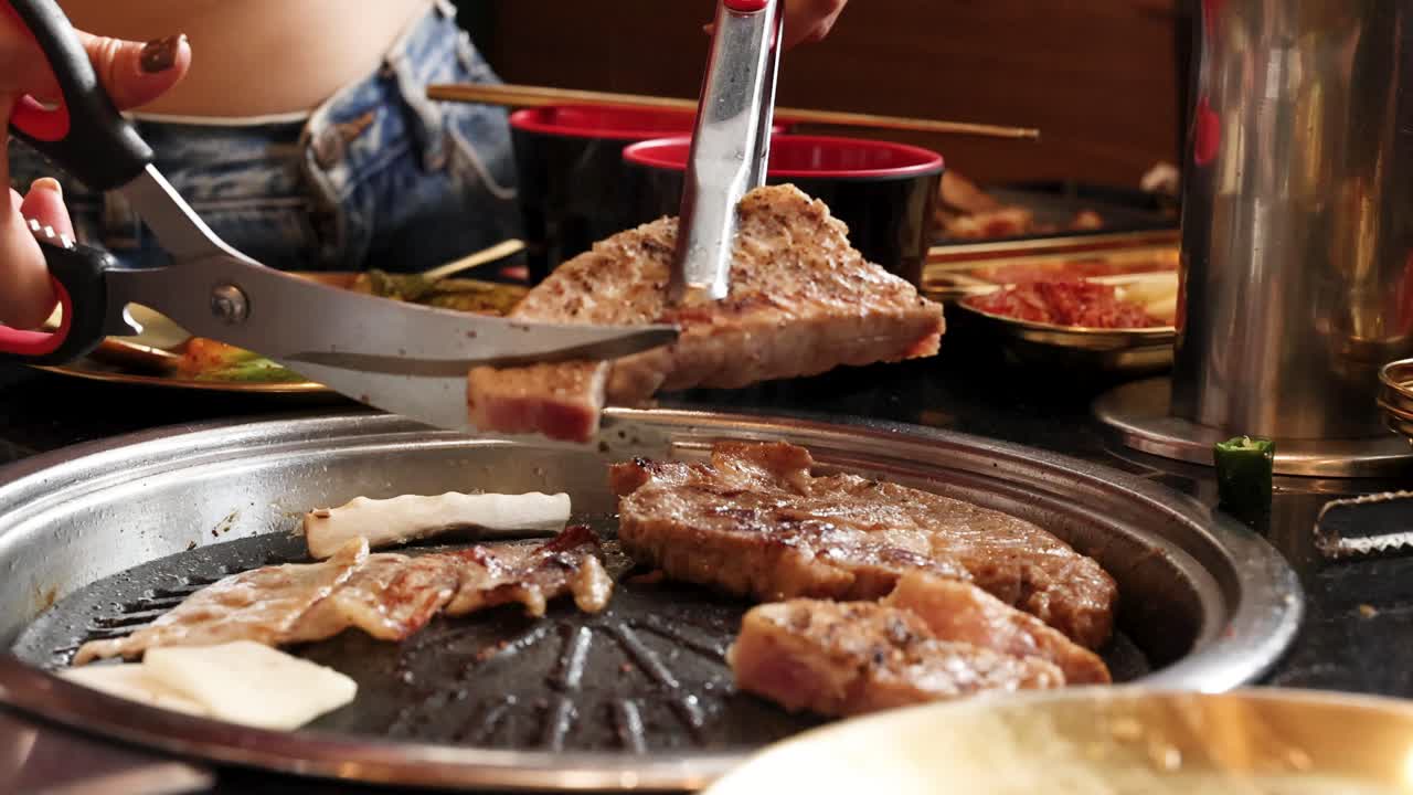 Person uses scissors to cut grilled pork belly on tabletop Korean barbecue under warm lighting