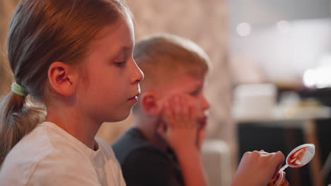 close up of young girl dipping fries into sauce container while boy beside her looks worried with hands on cheeks as they sit together indoors during meal time in soft evening light atmosphere