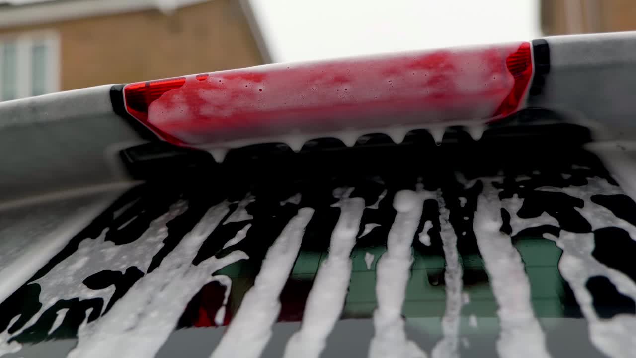 Close up orbiting shot of snow foam dripping from rear spoiler and brake light onto rear window of car