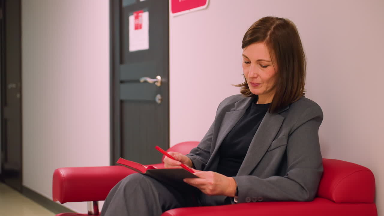 Businesswoman sitting on red couch in modern office corridor, writing in notebook with red pen. Focused on note-taking, relaxed atmosphere, professional work in a contemporary environment