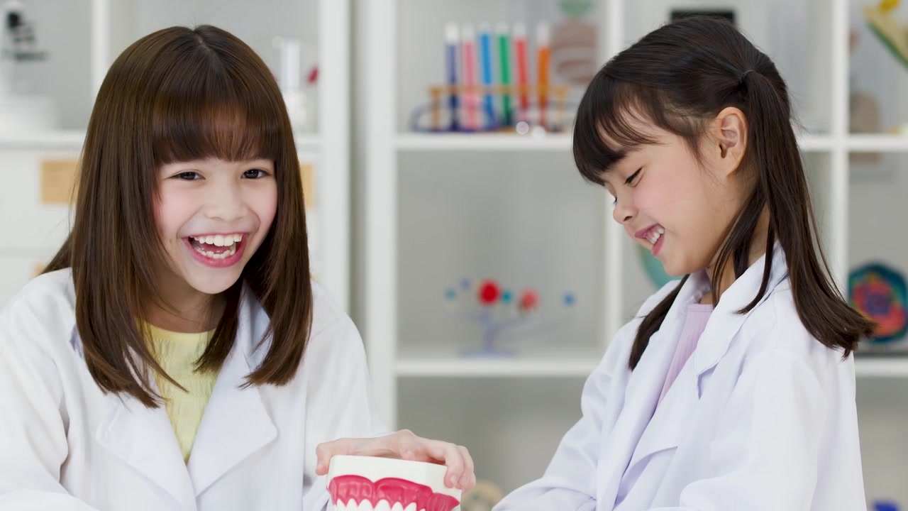 Hai cô gái trẻ mặc áo khoác phòng thí nghiệm màu trắng đang nghiên cứu mô hình răng trong một lớp học sáng sủa