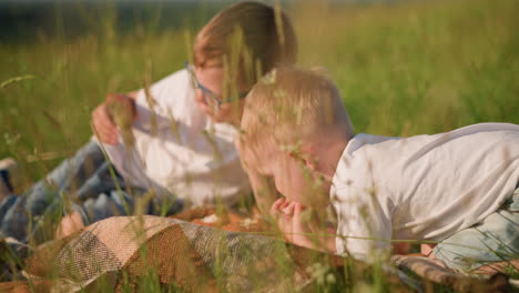 close-up of two young boys in white tops, one lying down and the other bent over on a checkered scarf in a grassy field, as they enjoy eating bread together. natural moment of childhood bonding