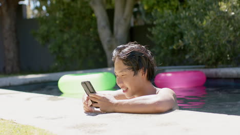 Estudiante-Universitario-Asiático-Sentado-Junto-A-La-Piscina,-Mirando-El-Teléfono-Inteligente