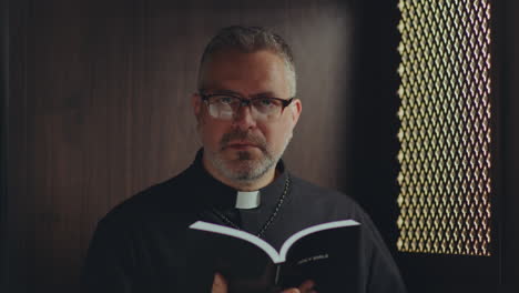 portrait of catholic priest holding bible in confessional booth