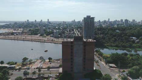 Experimente-El-Paisaje-Urbano-Junto-A-Bulliciosos-Sitios-De-Construcción,-Con-El-Telón-De-Fondo-De-Un-Río-Sereno-Y-árboles-Verdes,-Todo-Capturado-En-Impresionantes-Imágenes-Tomadas-Con-Drones-En-Un-Día-Soleado.