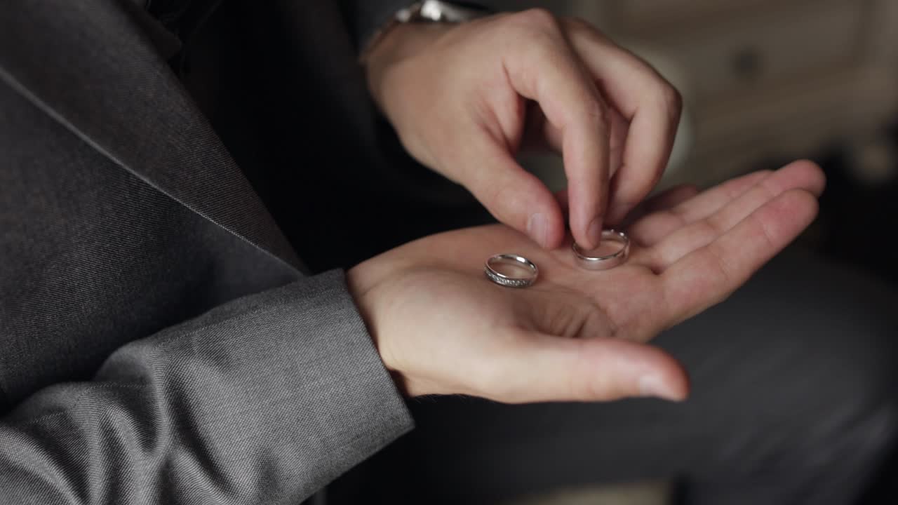 Groom in gray jacket holding wedding rings on palm of his hand, man touches the rings, slow motion