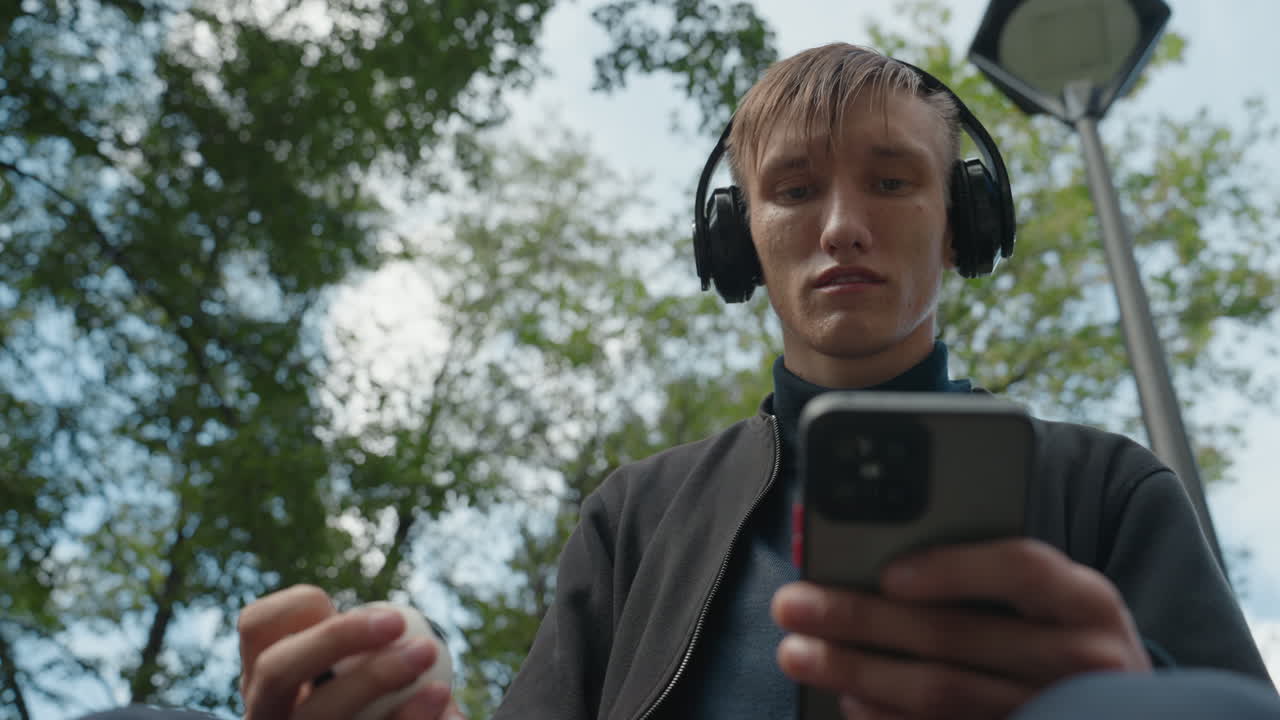 Giovane uomo bianco all'aperto che tiene in mano un telefono, indossa cuffie over-ear e controlla un piccolo auricolare, lampione e chioma di albero sopra la testa, espressione concentrata, product tester che esamina il prototipo audio sul telefono