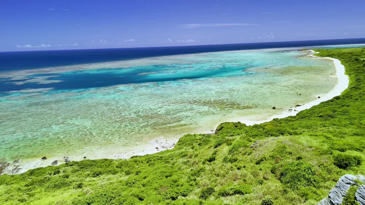Et fejende droneoptagelse fra frodige grønne bjergrygge, der afslører et stort, krystalklart turkist koralrev og en hvid sandstrand under en strålende blå himmel i Ishigaki, Japan.