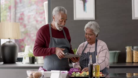 Senior-biracial-couple-cooking-dinner-and-using-tablet-in-kitchen,-unaltered,-in-slow-motion