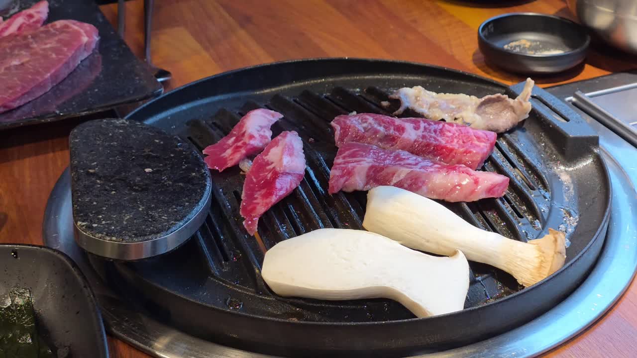 Hand places marbled beef slices and king oyster mushrooms onto a hot tabletop grill at a Korean BBQ restaurant.