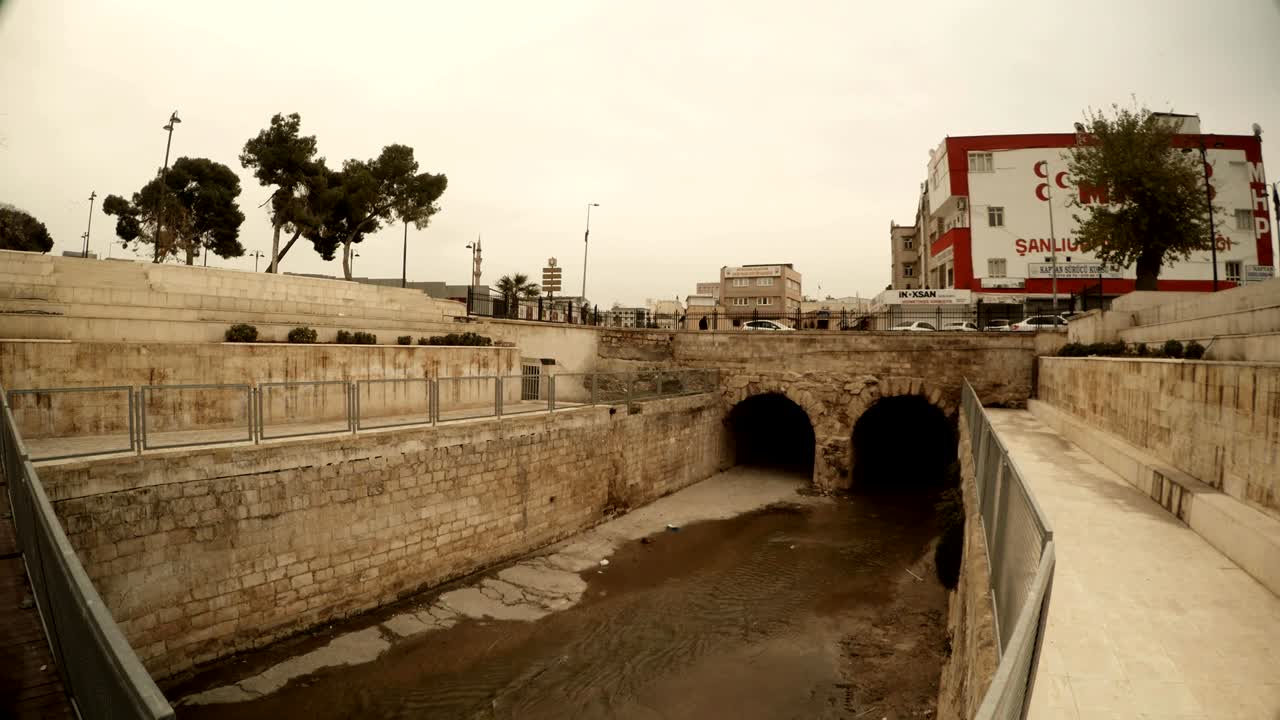 Vecchio ponte di pietra sotto un antico canale, auto che attraversano, Sanliurfa, giornata invernale nuvolosa.