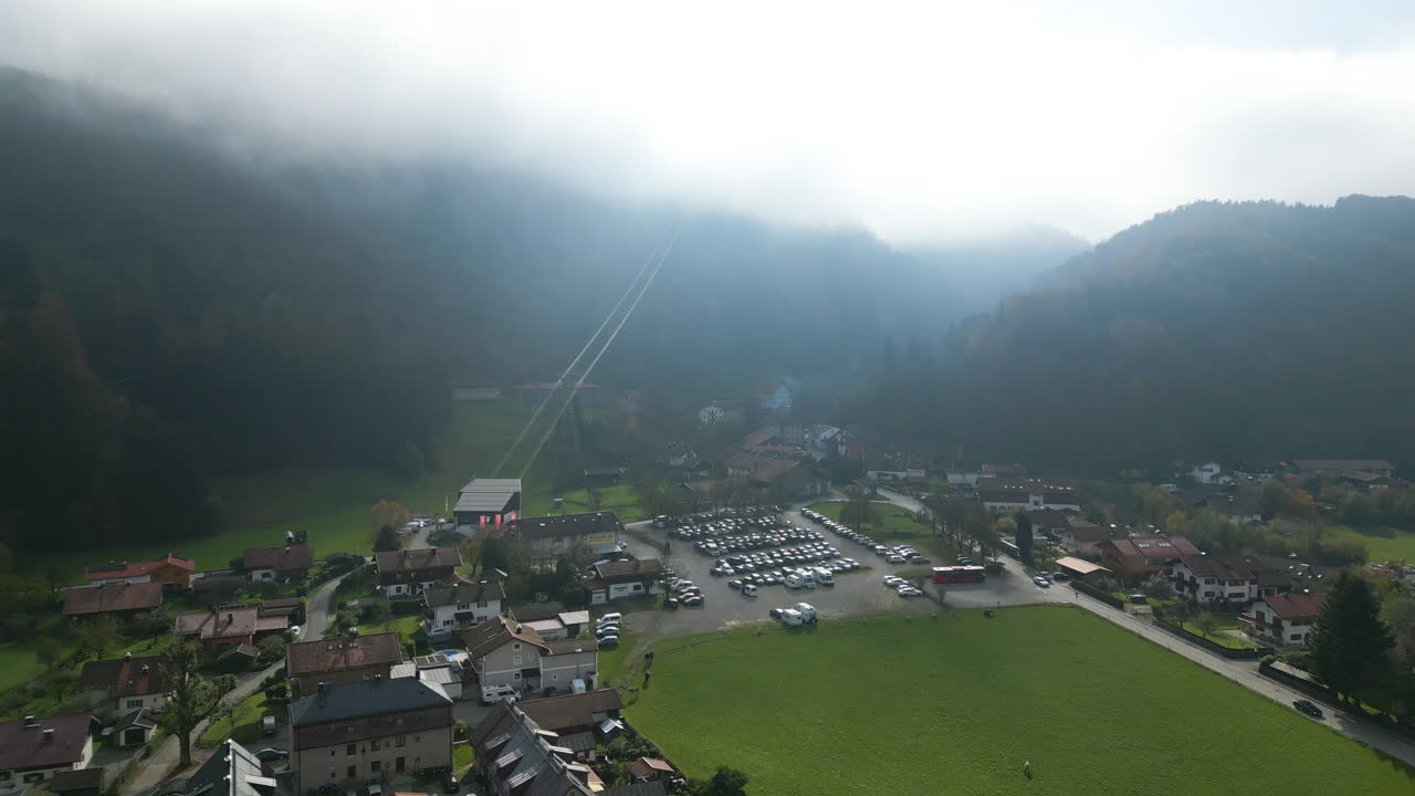 Scenic aerial shot of Bergen, a picturesque Bavarian village with clustered homes, green fields, and a cable car station, nestled below mist-covered mountains under a soft, diffused sky.