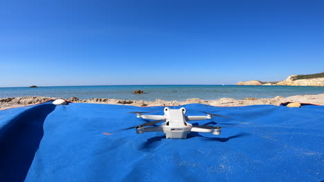 un dron comienza a hacer girar sus hélices sobre una toalla, en una playa griega para despegar