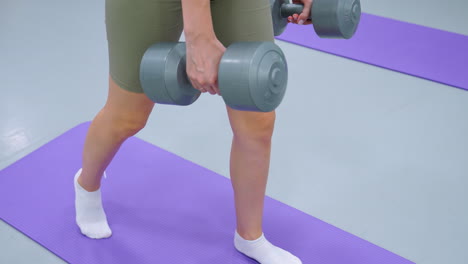 primer plano de una mujer ajustando la postura mientras levanta pesas en una alfombra de ejercicio púrpura, centrándose en la parte inferior del cuerpo y el agarre, enfatizando la fuerza, el equilibrio y el movimiento controlado en la rutina de entrenamiento de fitness