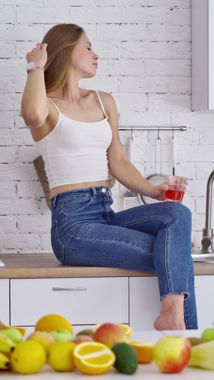Beautiful woman on the table with juice. Young model in jeans sitting on the table at home and drinking fresh juice. Sexy female in the modern kitchen. Vertical video