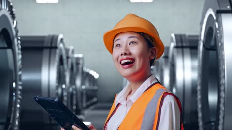 close up side view of asian female engineer with safety helmet looking at the tablet in her hand and looking around while standing in metal factory