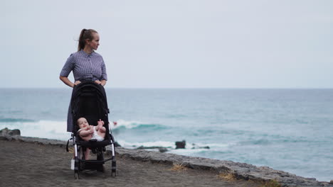 This-sweet-young-family's-dynamic-shines-as-a-young-mother-interacts-with-her-baby-in-the-stroller-against-the-ocean's-backdrop,-demonstrating-her-affection-and-devotion