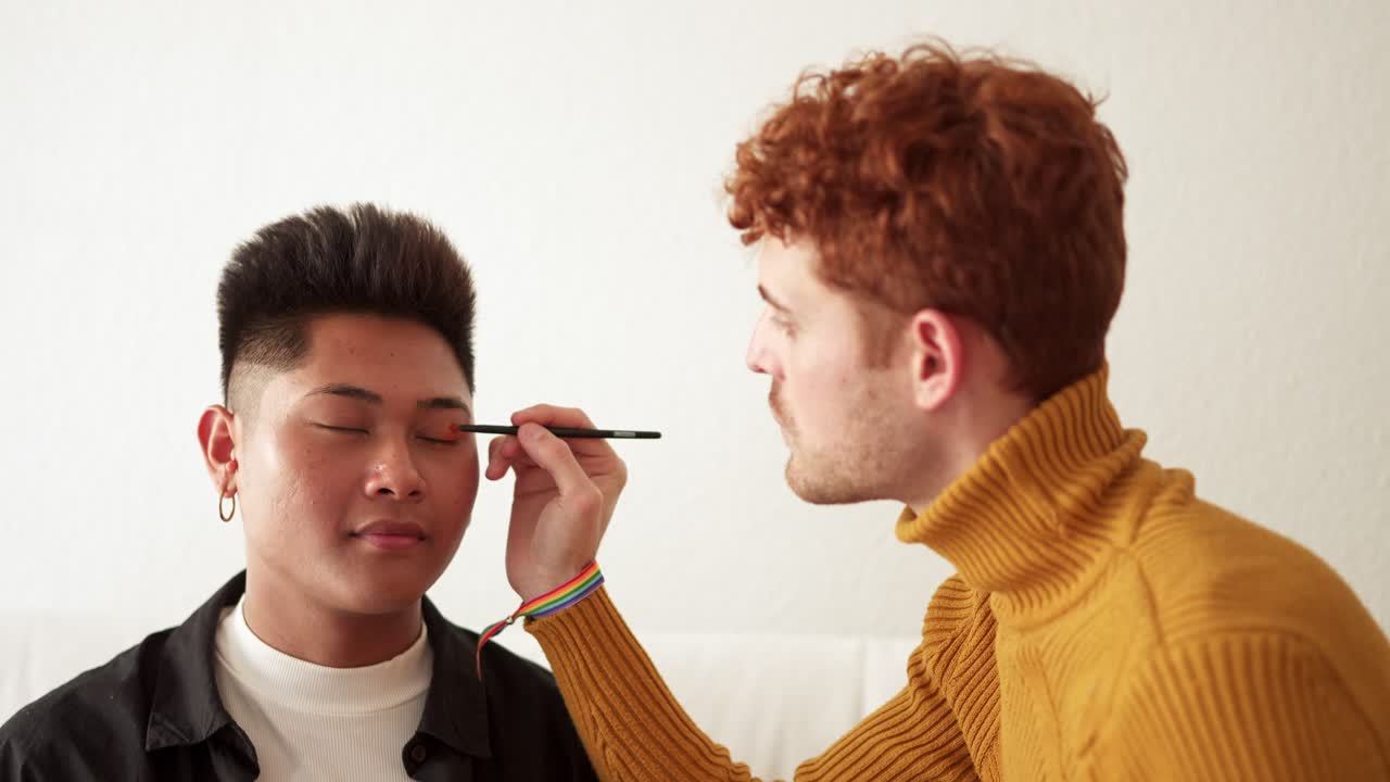 A person applies eyeshadow to another person, with a rainbow wristband visible on the makeup artist's wrist
