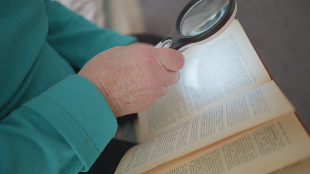 Senior woman reading book with magnifying glass, focused on text with clear details. Soft indoor lighting highlighting pages, showing care and attention to reading. Elderly care, senior leisure