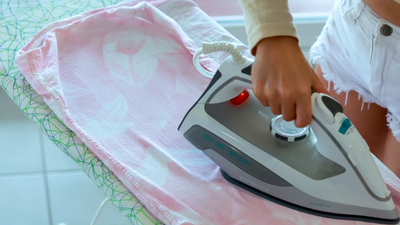A woman in white shorts irons a pink shirt on an ironing board, using a steam iron in a sunlit, modern interior with tiled flooring