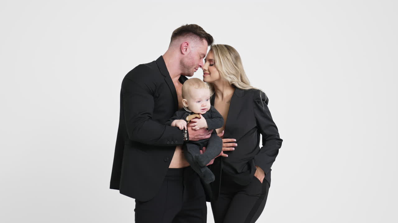 Caucasian couple wearing black suits with their infant kid. Happy family of three studio footage. White backdrop.