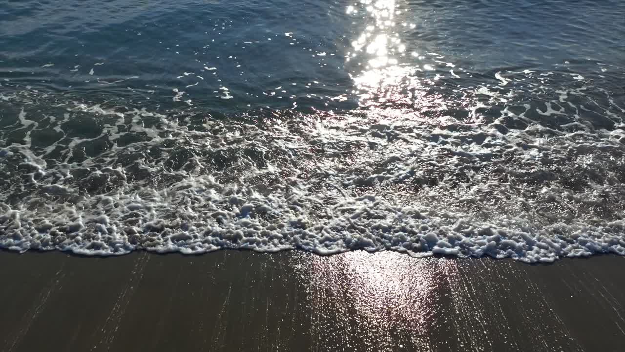Slow motion aerial pullback of stunning, turquoise ocean waves crashing against sandy beach shoreline revealing sunlit horizon and boat in background