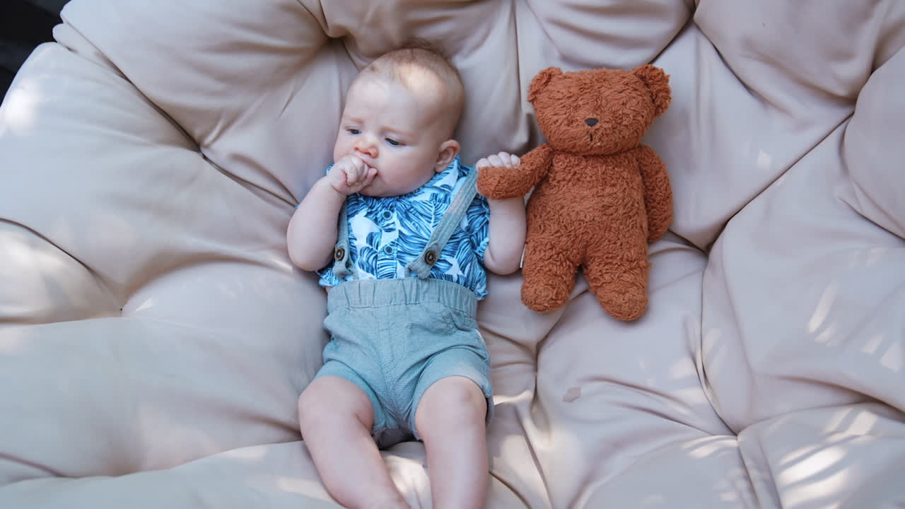 Adorable baby boy wearing shirt and shorts lies on the soft mattress. Infant sucking his fist and holding a teddy. Top view.