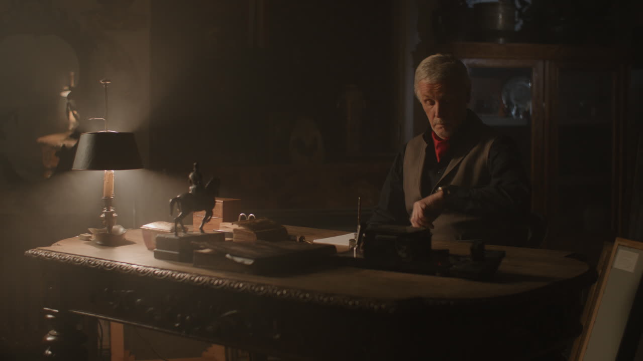 Old passionate man sits down to his desk, put on the glasses and writes a letter