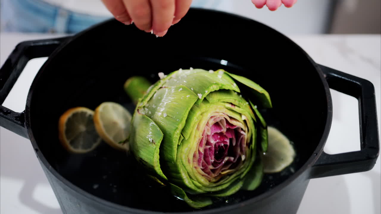 Woman pouring salt over an artichoke in a pot with water and lemons