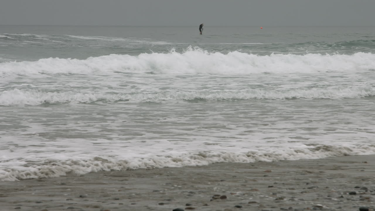 A lone paddle boarder rides the waves in Encinitas, California during an overcast morning.