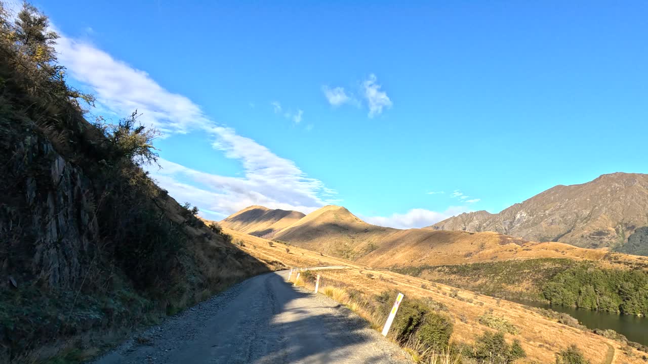 Vehicle travels gravel road beside lake, golden hills, and mountains under clear blue sky, daytime