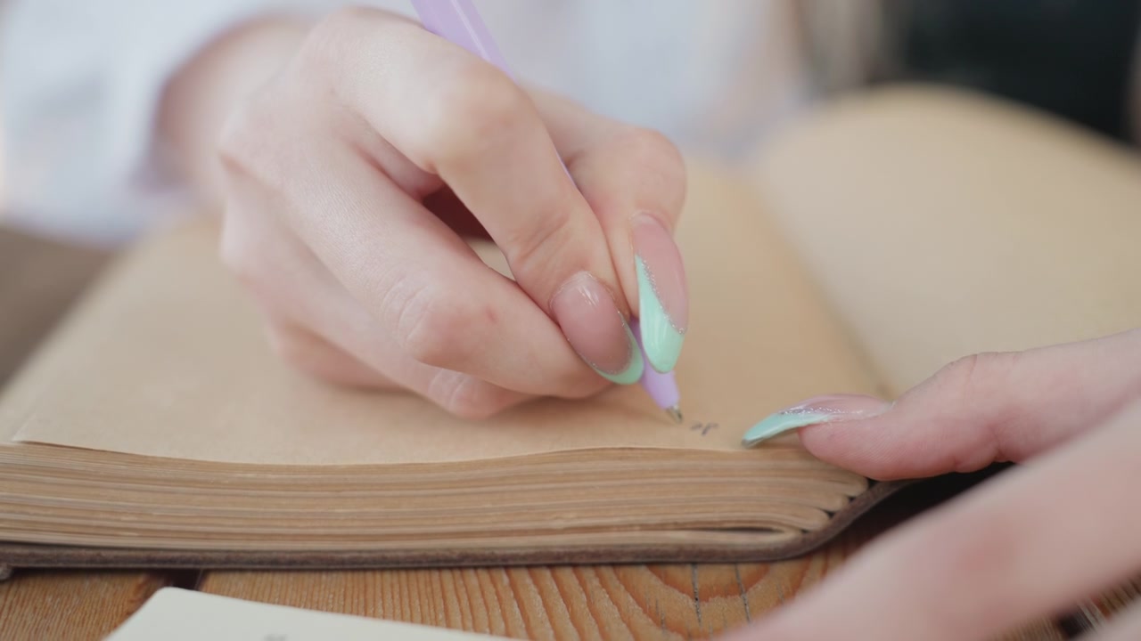 Close up of woman hand with pastel green nails writing in open notebook using purple pen. scene shows texture detail of paper and wood soft lighting and calm expression