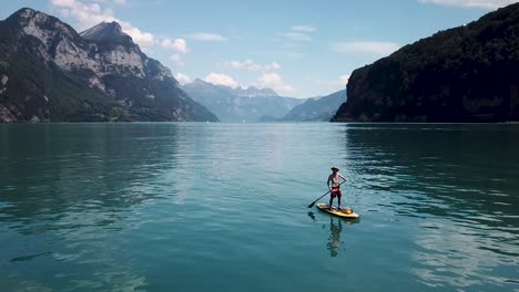 a white male is holding up his paddle on a stand up paddle, sup, the drone flies up to show him in the middle of a lake in switzerland